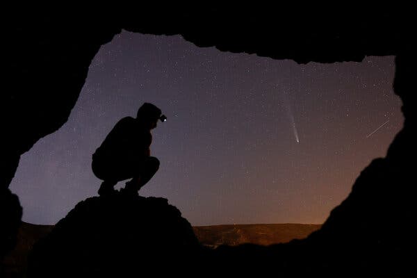 A person crouching at the entrance of a cave, in silhouette, to watch a comet in the night sky.