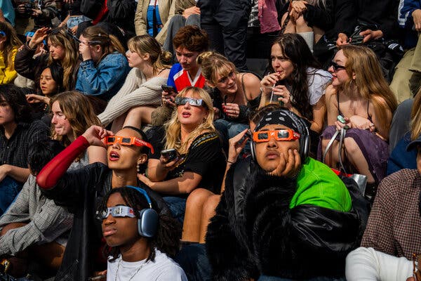 A crowd of young people with eclipse glasses sit and watch the sky.