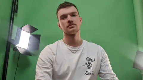 John O'Reilly John O'Reilly looking at the camera with a green screen behind him, wearing a white top and necklace.