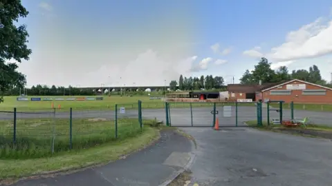 Google Google street view of the West Bank Bears ground in Widnes. There is a club house with a pitch in the background, along with advertisement boards. There is a green wire fence and a gate in the foreground.