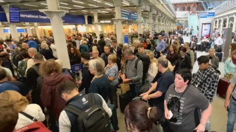 BBC A crowd of people at London St Pancras station in London. They're carrying rucksacks or suitcases as they wait to go through departures and board a Eurostar train across the Channel.