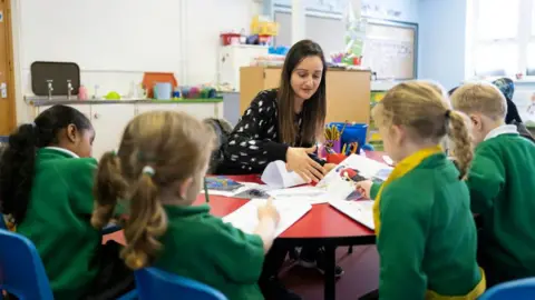 Getty Images A primary school teacher sitting with a group of young children in green school jumpers at a table in a classroom.