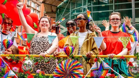 Getty Images Five people pictured behind a barrier at Manchester Pride. They are waving pride flags smiling and waving. The barrier is decorated with various rainbow flags and disco balls.