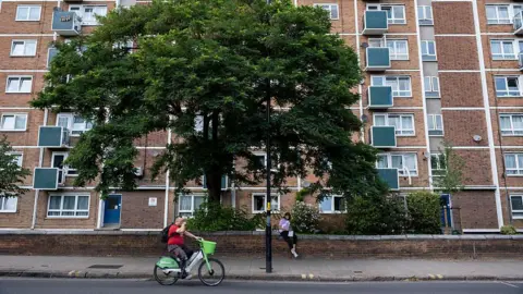 In Pictures via Getty Images A block of social housing flats viewed from the road, with a man cycling in front of them and a large leafy tree behind him, in front of the flats.