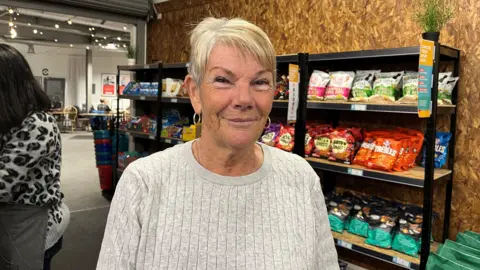 Kelly Foran/BBC Kathleen Roper, who has short blonde hair and wears a light grey jumper, smiles at the camera against a backdrop of a shelf contained large bags of crisps. 