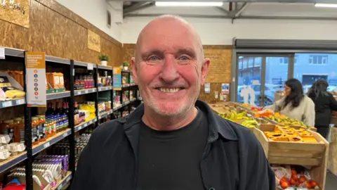 Kelly Foran/BBC Andy Hawthorne, who has a bald head and light grey stubble, beams at the camera inside a shop stocked with groceries. 